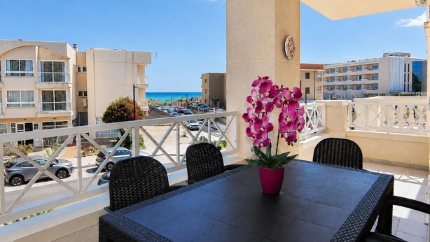 Varanda com mesa e cadeiras, uma orquídea em vaso e vista para a praia e edifícios.