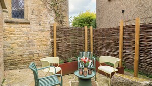 Un bâtiment en pierre avec une clôture en bois, des sièges extérieurs comprenant des chaises et une table, et une fenêtre dotée d’un grillage métallique.