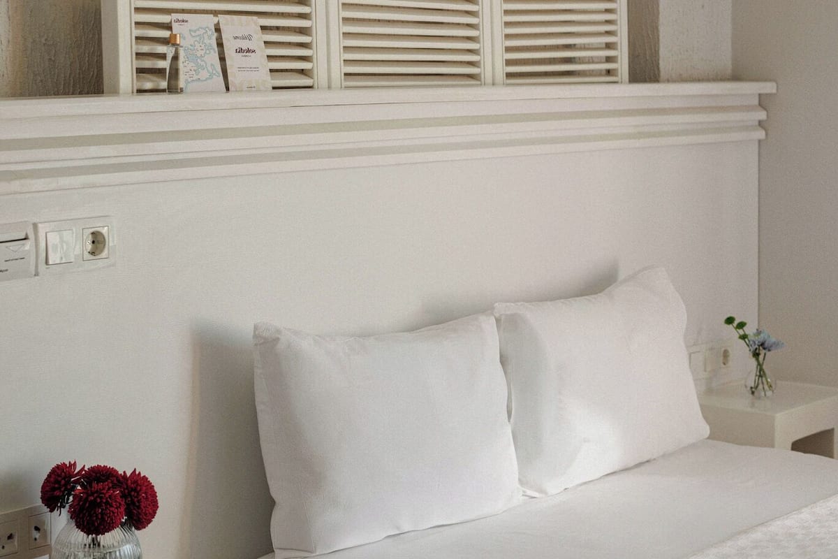 A bed with white bedding, a nightstand with a vase of flowers, and a window with blinds.