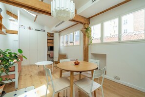 Ein Zimmer mit Holzboden, weißen Wänden, einem runden Holztisch mit Stühlen, einer hängenden Pflanze und großen Fenstern.