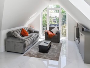 Un loft moderno con techo alto, un sofá gris con cojines naranjas, una mesa de centro negra y vistas a un paisaje verde a través de un ventanal grande.