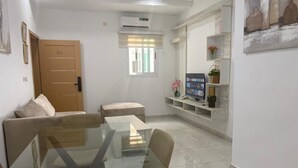 A modern living room with a glass dining table, a sofa, a TV, and a shelf with decorative items.