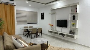 A modern living room with a sofa, a dining table with chairs, a TV mounted on a wall, and a small shelf with decorative items.