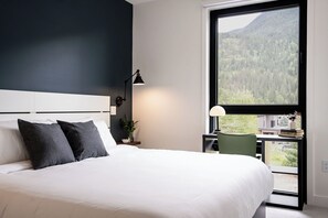 Ein Schlafzimmer mit einem großen Bett, einem Schreibtisch mit Stuhl, einem Fenster mit Blick auf einen Wald und einer Lampe auf dem Nachttisch.