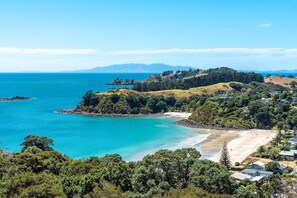 A coastal landscape with a beach, dense greenery, and distant mountains. - Miro Ridge in Palm Beach - Miro Ridge is a spacious elevated holiday home overlooking Palm Beach with sea views and generous indoor-outdoor living. Large sliding doors open to an expansive deck, while the open plan lounge and entertainer’s kitchen cr (Waiheke Island)