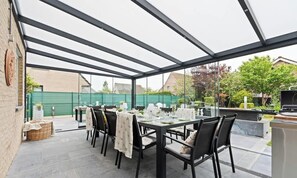 Een overdekte eethoek buiten met tafels voor twee personen, een barbecue en uitzicht op de omliggende tuin en gebouwen.