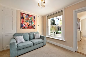 A room with a grey sofa, a colorful abstract painting, a window with a view of outdoor furniture, and a white door. - Finest Retreats | Yew Cottage (Tunbridge Wells)