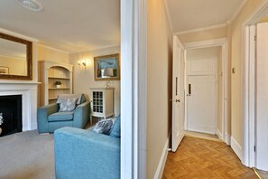 A living room with a fireplace, a blue sofa, a white cabinet, and a hallway leading to a door. - Finest Retreats | Yew Cottage (Tunbridge Wells)