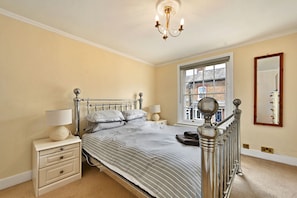 A bedroom with a bed, bedside tables, a lamp, a mirror, and a window with a view of buildings. - Finest Retreats | Yew Cottage (Tunbridge Wells)