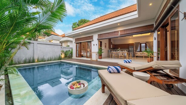 Uma área de piscina moderna com espreguiçadeiras, espaço para refeições e cozinha visível através do ambiente aberto.