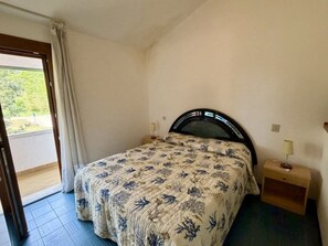 Ein Schlafzimmer mit Bett, Nachttisch, Lampe, einem Fenster mit Vorhängen und Blick ins Grüne.