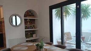 A dining area with a round table, chairs, a chandelier, a mirror, shelves with decorative items, and a view of the outdoors.