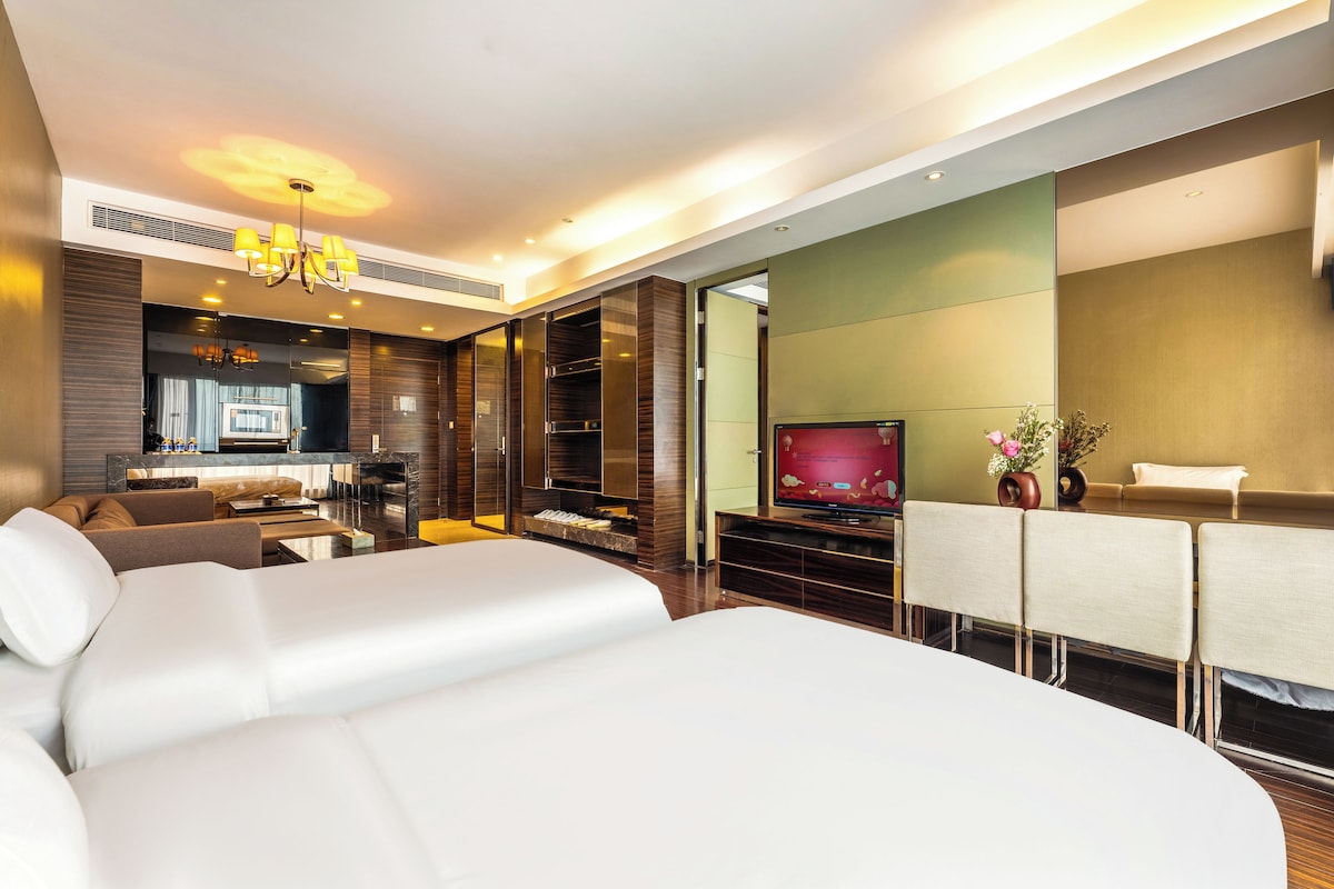 Una habitación de hotel moderna con una cama grande, televisión, zona de comedor y cocina al fondo.