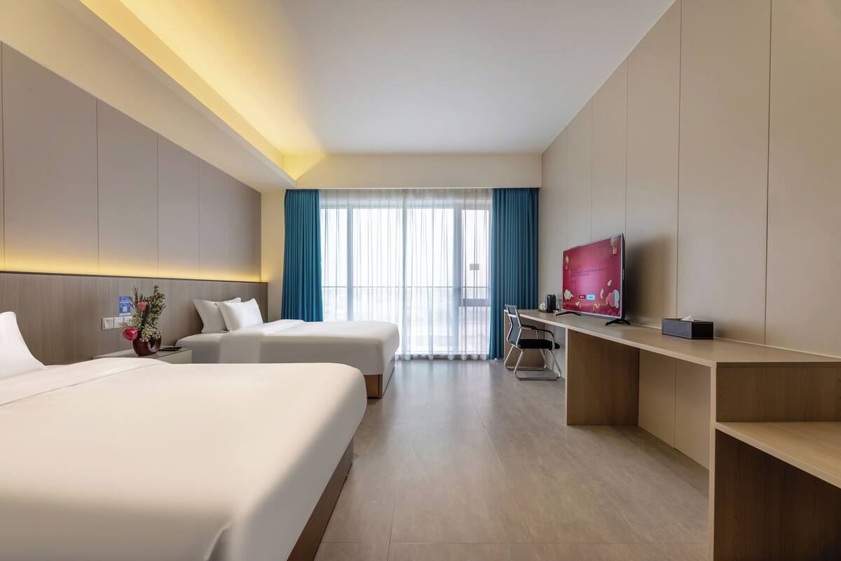 Habitación de hotel con dos camas, un escritorio, un televisor y un ventanal con cortinas.