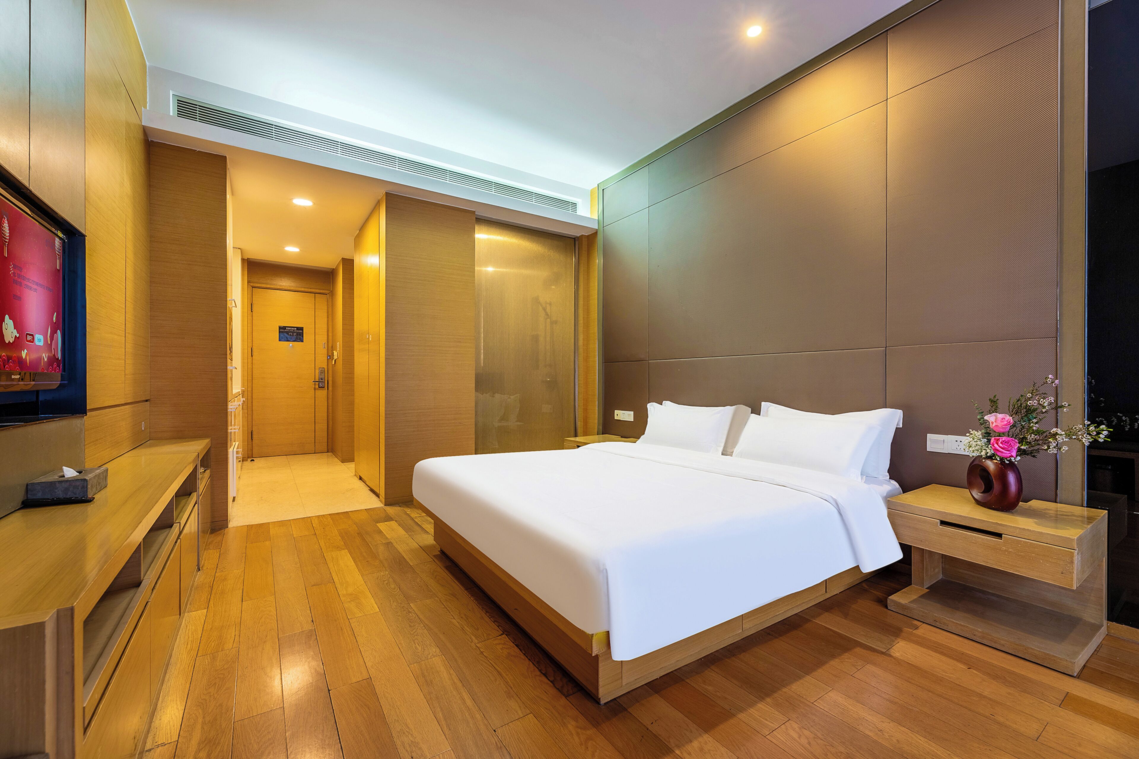 A modern hotel room with a large bed, wooden flooring, a nightstand with a vase of flowers, and a television on the left.