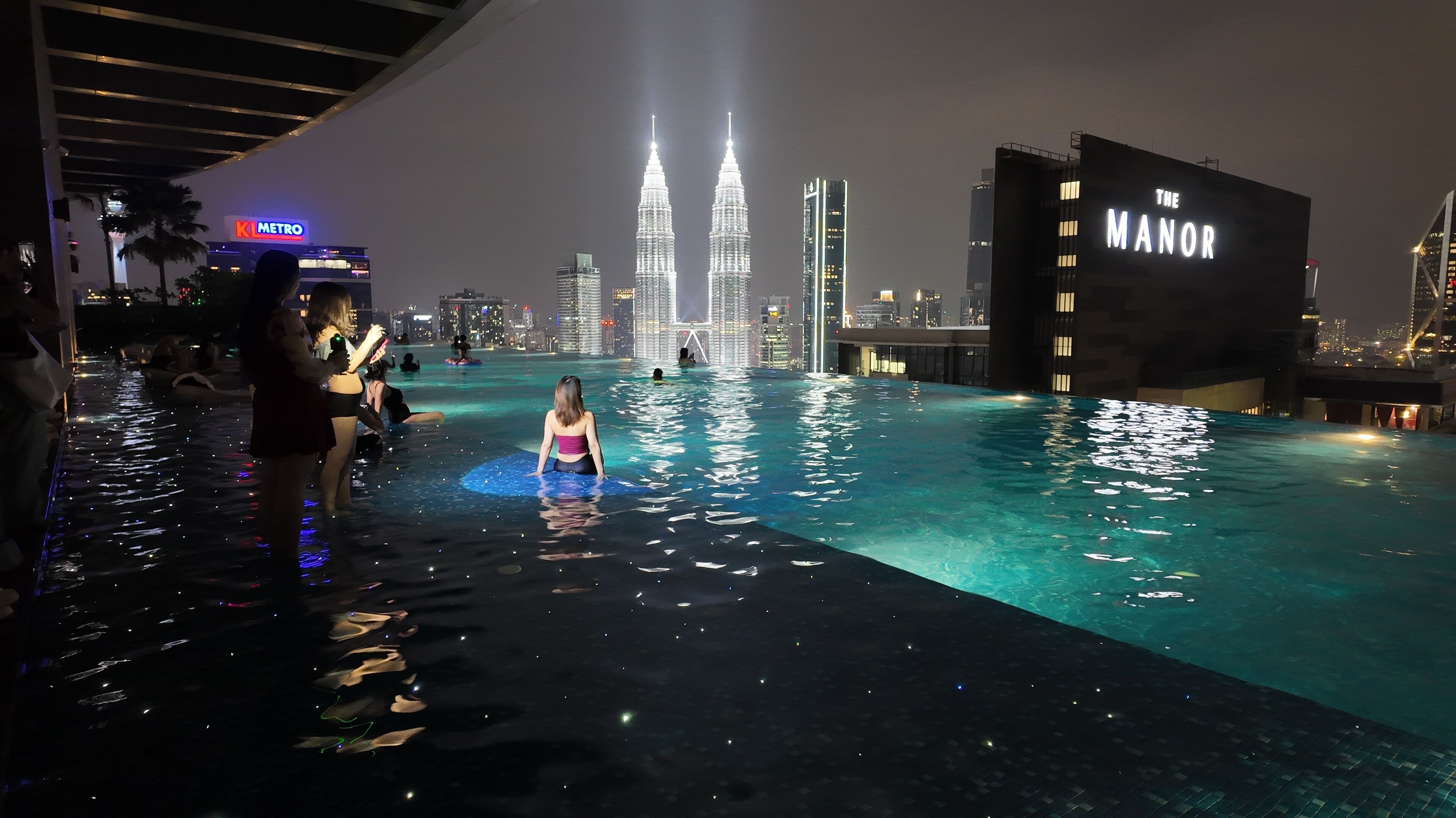 An infinity pool with a city skyline view, including the Petronas Towers, at night.