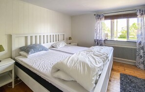 Uma cama de casal com roupa de cama branca, uma mesa de cabeceira com abajur, uma janela com cortinas e um radiador.