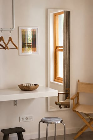 A corner room with a white wall, a window with curtains, a wooden chair, a stool, a shelf with a bowl, and a framed abstract painting.
