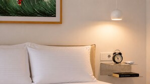 A neatly made bed with white linens, a bedside table with a classic alarm clock, a small shelf with a book, and a framed artwork above the bed. - Ayvalık Center Modern Detached House in the City Center (Ayvalık)