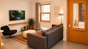 A modern living room with a grey sofa, a black chair, a small table, a potted plant, and a television on the wall. - Ayvalık Center Modern Detached House in the City Center (Ayvalık)