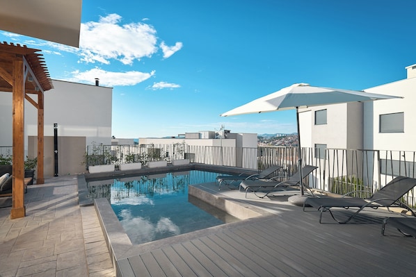 Ein Pool auf dem Dach mit Liegestühlen und Sonnenschirmen, umgeben von einer Holzterrasse und einem weißen Gebäude.