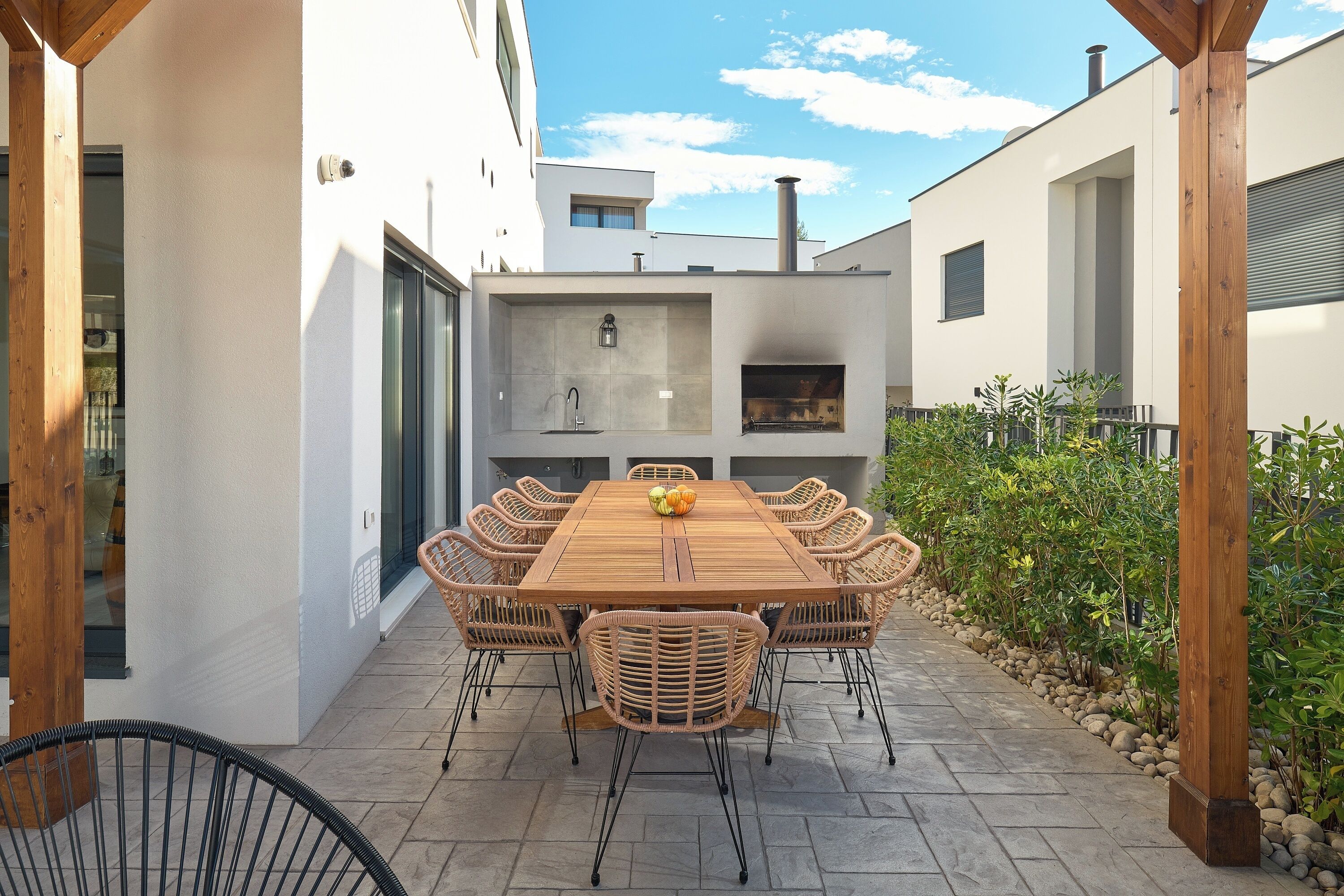An outdoor patio with a wooden table and wicker chairs, a built-in grill, and a stone-tiled floor.