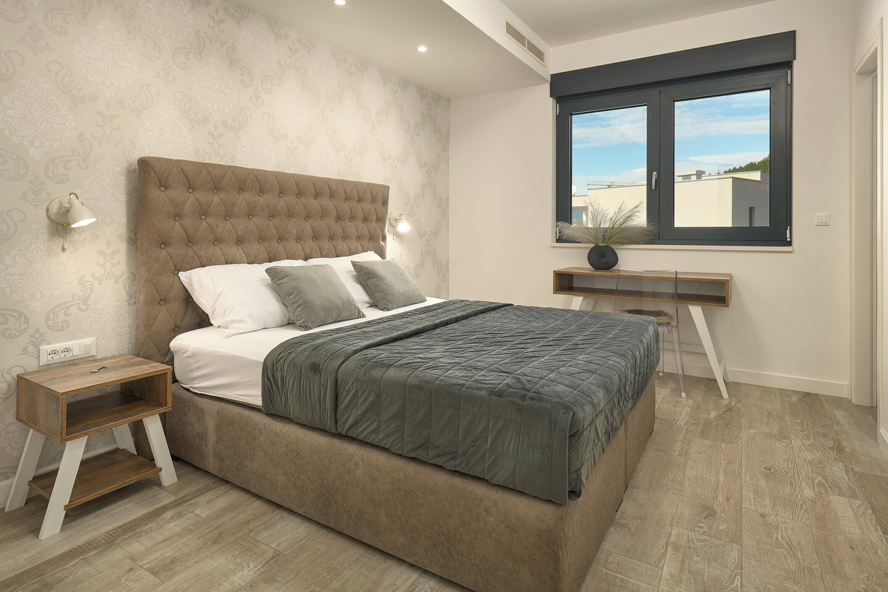Une chambre à coucher avec une tête de lit capitonnée, une table de chevet, un bureau et une fenêtre donnant sur un beau paysage.