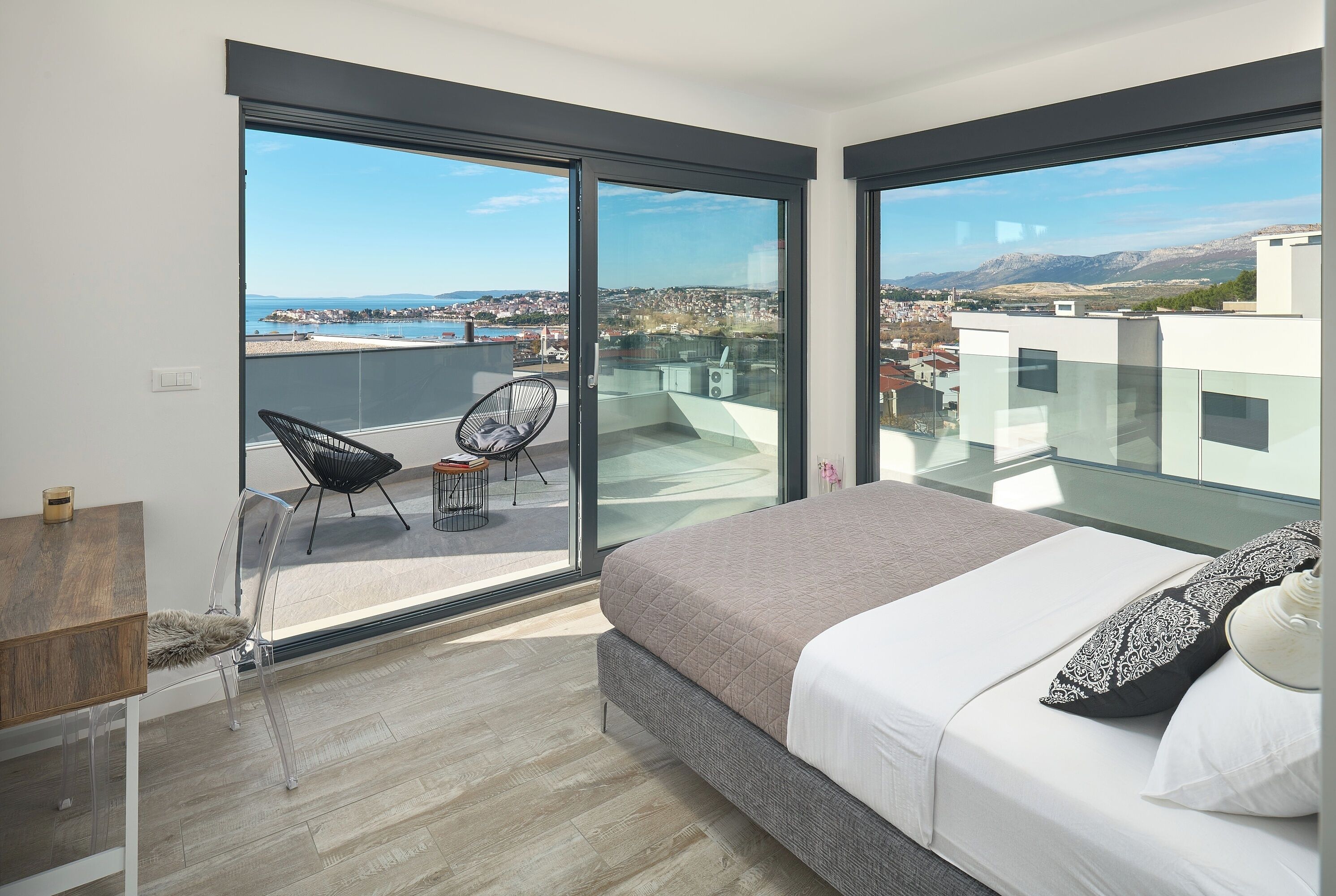 A bedroom with a large bed, a chair, a table, and a view of a city and mountains through large windows.
