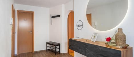 Un pasillo con una puerta de madera señalizada con 'WC', un pequeño banco negro, un espejo redondo y una consola de madera con un jarrón y un certificado enmarcado.
