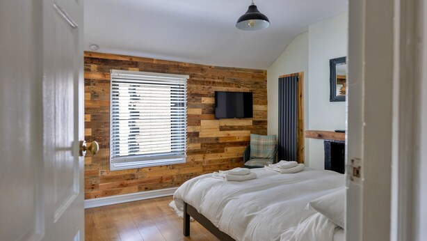 A bedroom with a wooden wall panel, a bed, a window with blinds, a chair, a TV, and a hanging light fixture. - Ty Pen Y Bryn, Castle Views (Conwy)