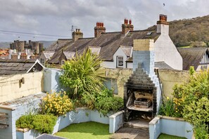Ein Innenhof mit Kamin aus Stein, ein kleiner Garten mit gelben Blumen und traditionelle Häuser mit geneigten Dächern.