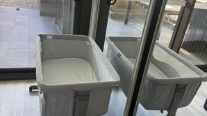 A room with two baby cribs against a glass wall, a wooden floor, and a view of a balcony.