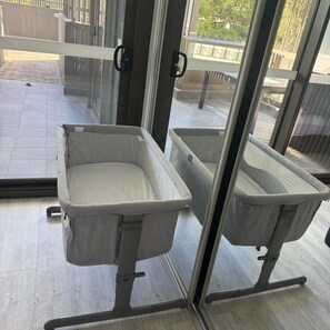 Ein Zimmer mit zwei Babybetten an einer Glaswand, Holzboden und Blick auf einen Balkon.
