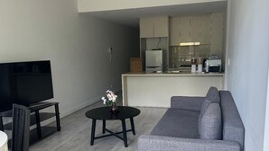 A living room with a gray sofa, a TV on a stand, a small round table with a vase, and a kitchenette in the background.