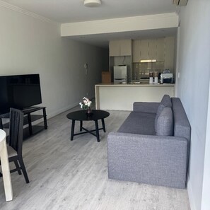 Ein Wohnzimmer mit grauem Sofa, einem Fernseher auf einem Schrank, einem kleinen runden Tisch mit einer Vase und einer Küchenzeile im Hintergrund.