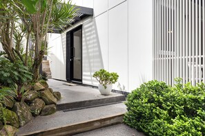 Ein modernes Gebäude mit einer Betontreppe, die zu einer schwarzen Tür führt, umgeben von Grünpflanzen und einem weißen Zaun.