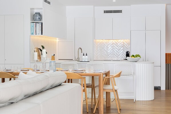 Una cocina moderna con armarios blancos, un comedor con sofá blanco, sillas de madera y una encimera blanca con salpicadero de espiga.