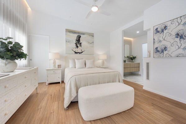 A bedroom with a bed, bedside tables, a nightstand, a large window with blinds, a ceiling fan, and artwork on the walls. - Absolute Beachfront - Seaside Serenity - sand dunes - Currimundi, Sunshine Coast (Currimundi)