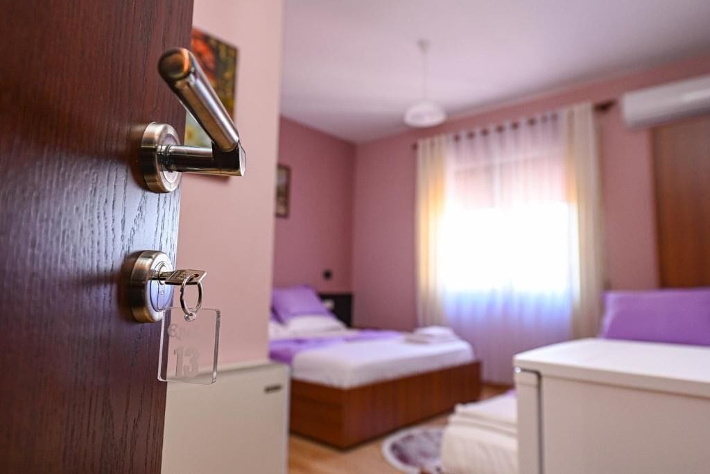 A hotel room with a wooden door, a bed with a headboard, a bedside table, and a window with curtains.