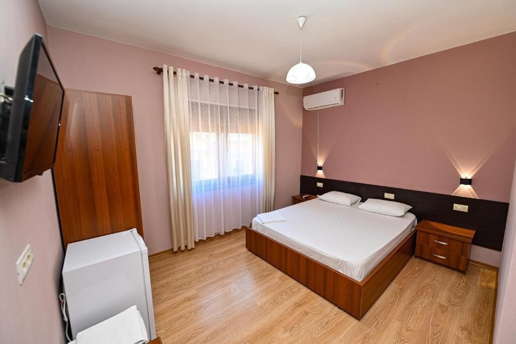 Une chambre à coucher avec un lit en bois, des rideaux blancs, un téléviseur à écran plat, un climatiseur et une lampe de chevet.