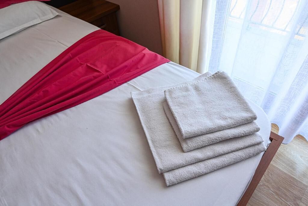 A neatly made bed with white and red linens, a stack of folded towels, and a wooden nightstand.