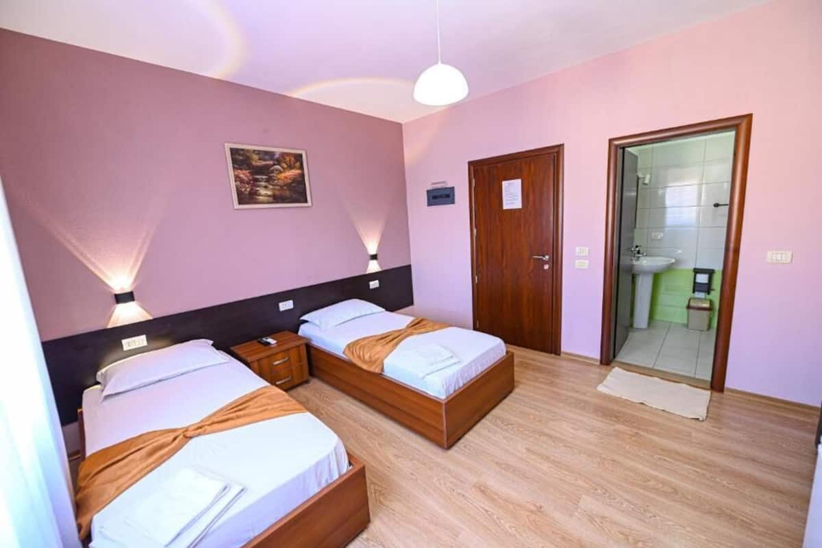 Habitación de hotel con dos camas, cabecera de madera, un cuadro en la pared, puerta al baño y una mesita de noche pequeña.