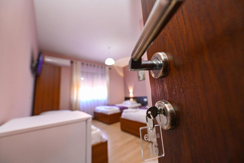 A hotel room with a wooden door, a bed, a bedside table, and a window with curtains.