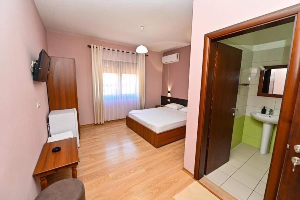 Habitación de hotel con cama, escritorio, silla, televisor, ventana con cortinas y baño con lavabo y espejo.