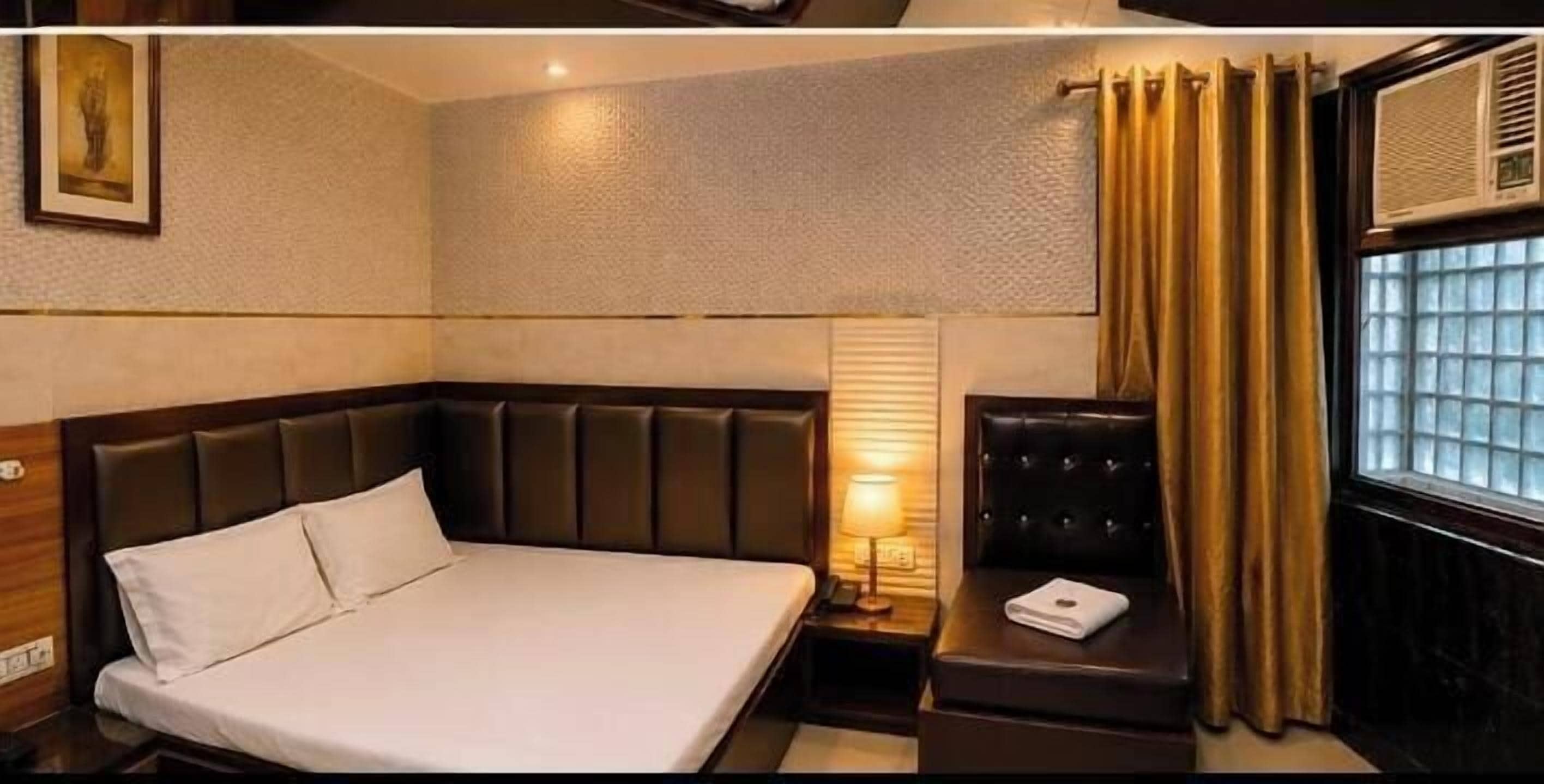 A hotel room with a large bed, a small seating area, a nightstand with a lamp, a window with curtains, and a wall-mounted air conditioner.