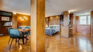 Una habitación amplia con piso de madera, un comedor con sillas, una cama con almohadones y estanterías empotradas de madera.