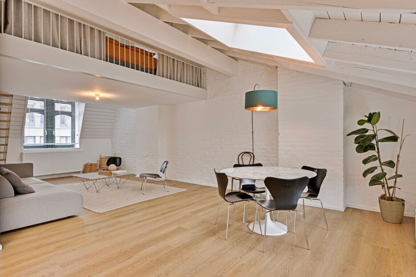 A spacious loft apartment with a high ceiling, a dining area with a round table and chairs, a sofa, a small coffee table, and a potted plant.