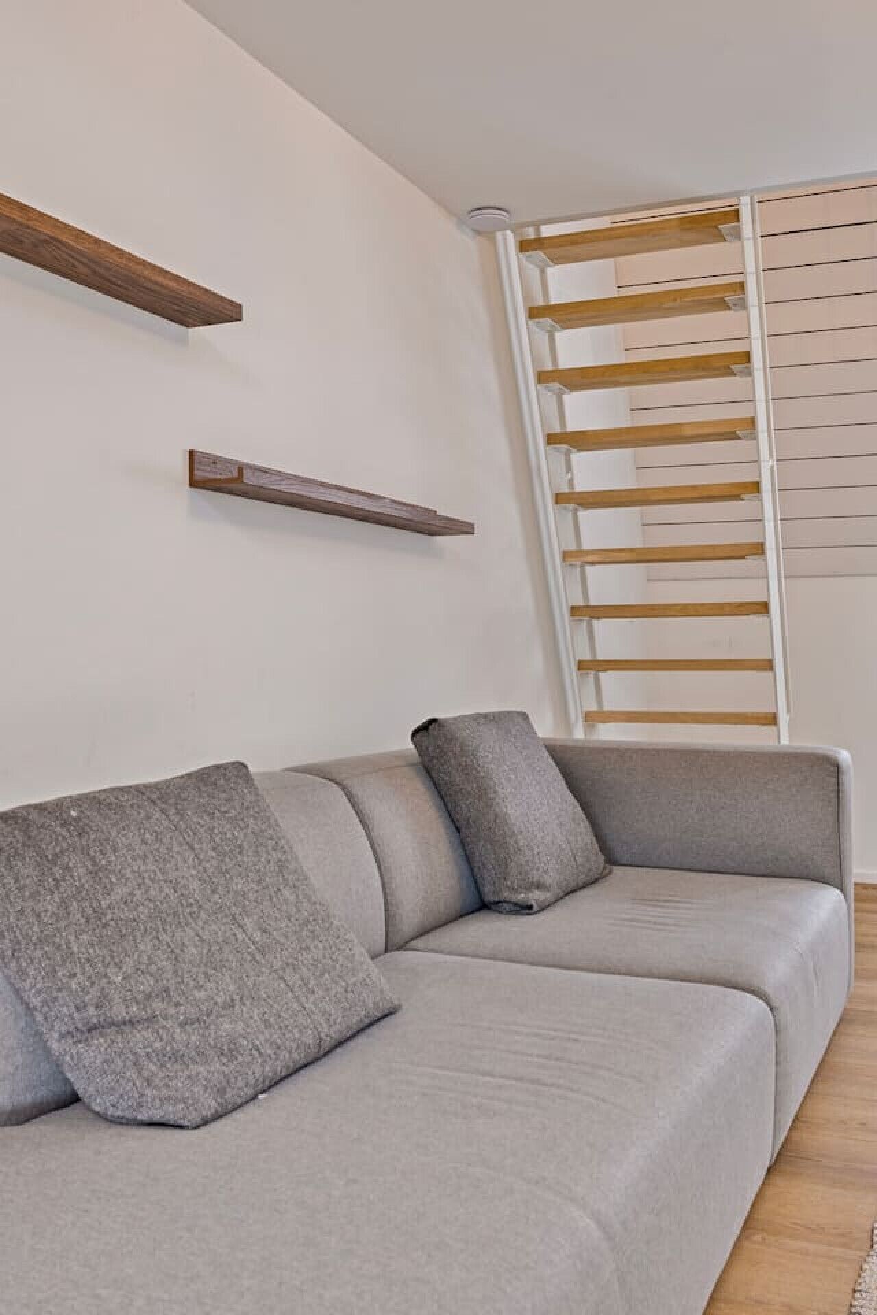 Un canapé gris avec deux coussins, une étagère en bois et un mur blanc avec une fenêtre.