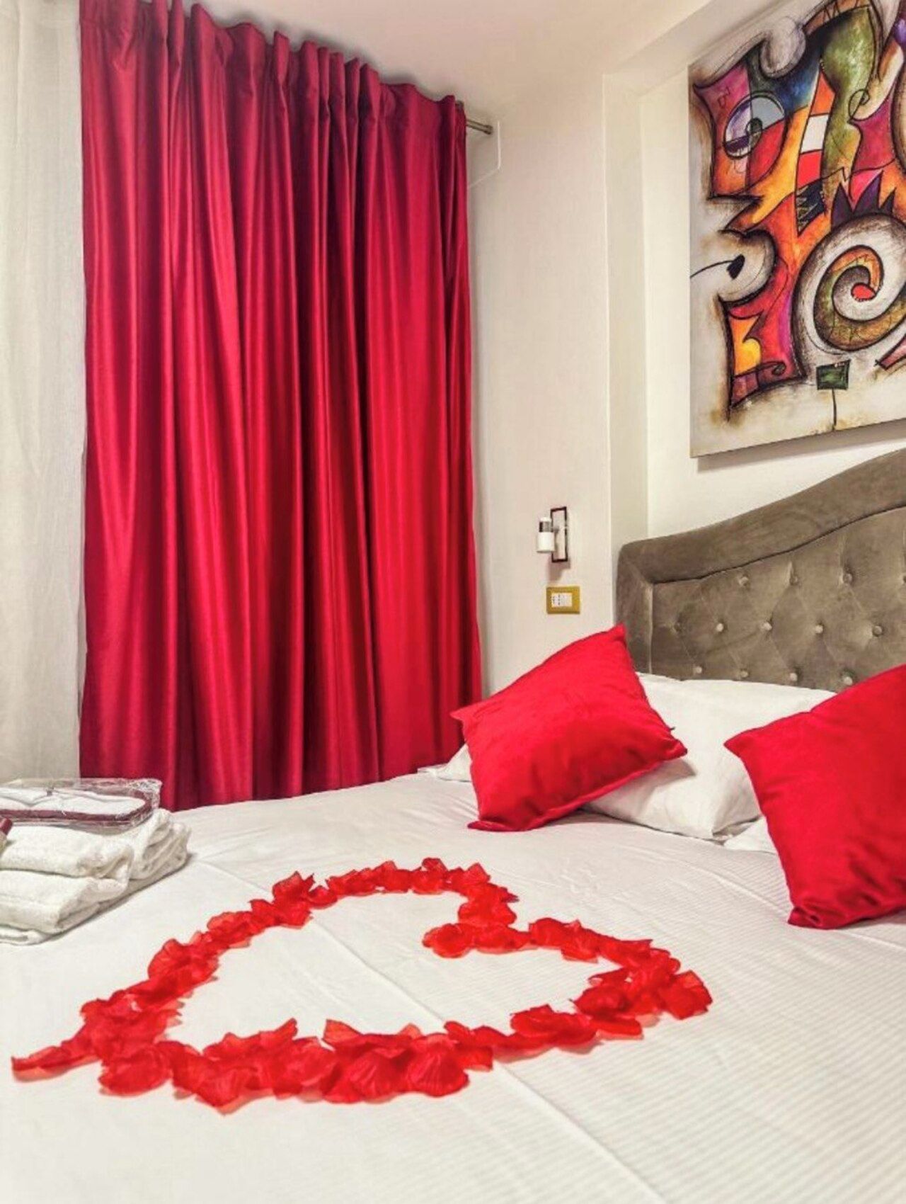 Habitación de hotel con cama, cortinas rojas, un cuadro abstracto de colores y una rosa roja decorando la cama.