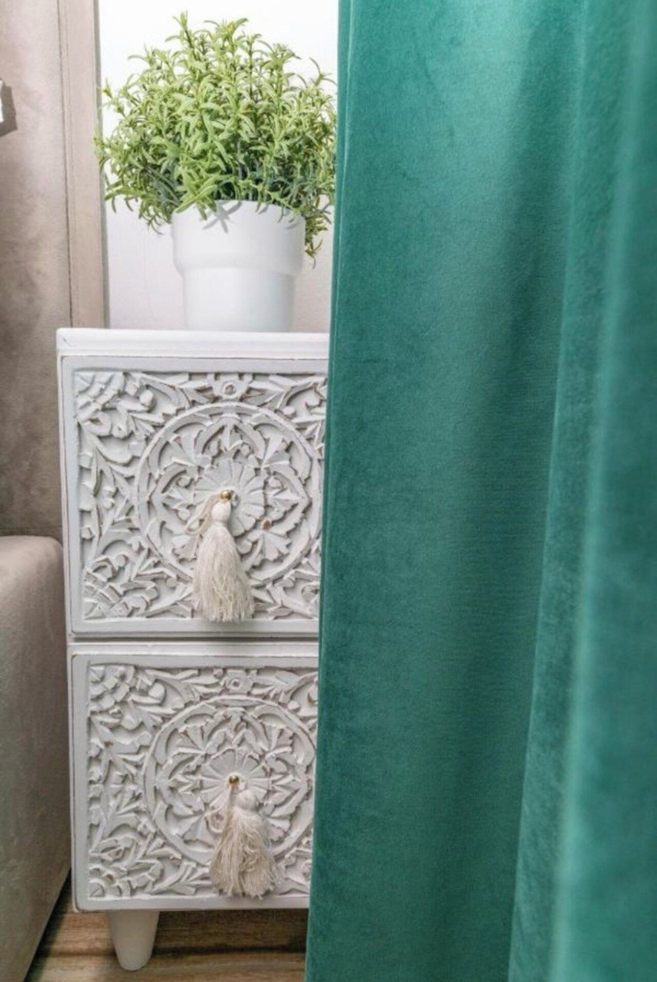 Une commode blanche ornée de motifs complexes, une plante en pot posée sur le dessus et un rideau turquoise à moitié tiré.
